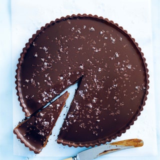 A salted chocolate caramel tart garnished with flaky sea salt with a cut slice and a knife next to it.